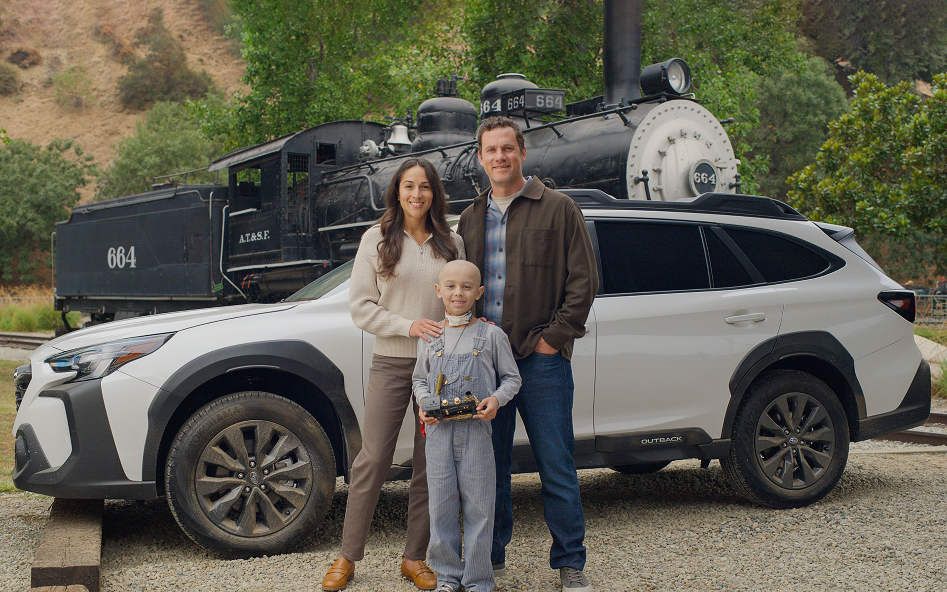Boy holding toy train standing with parents in front of a Subaru Outback and train.