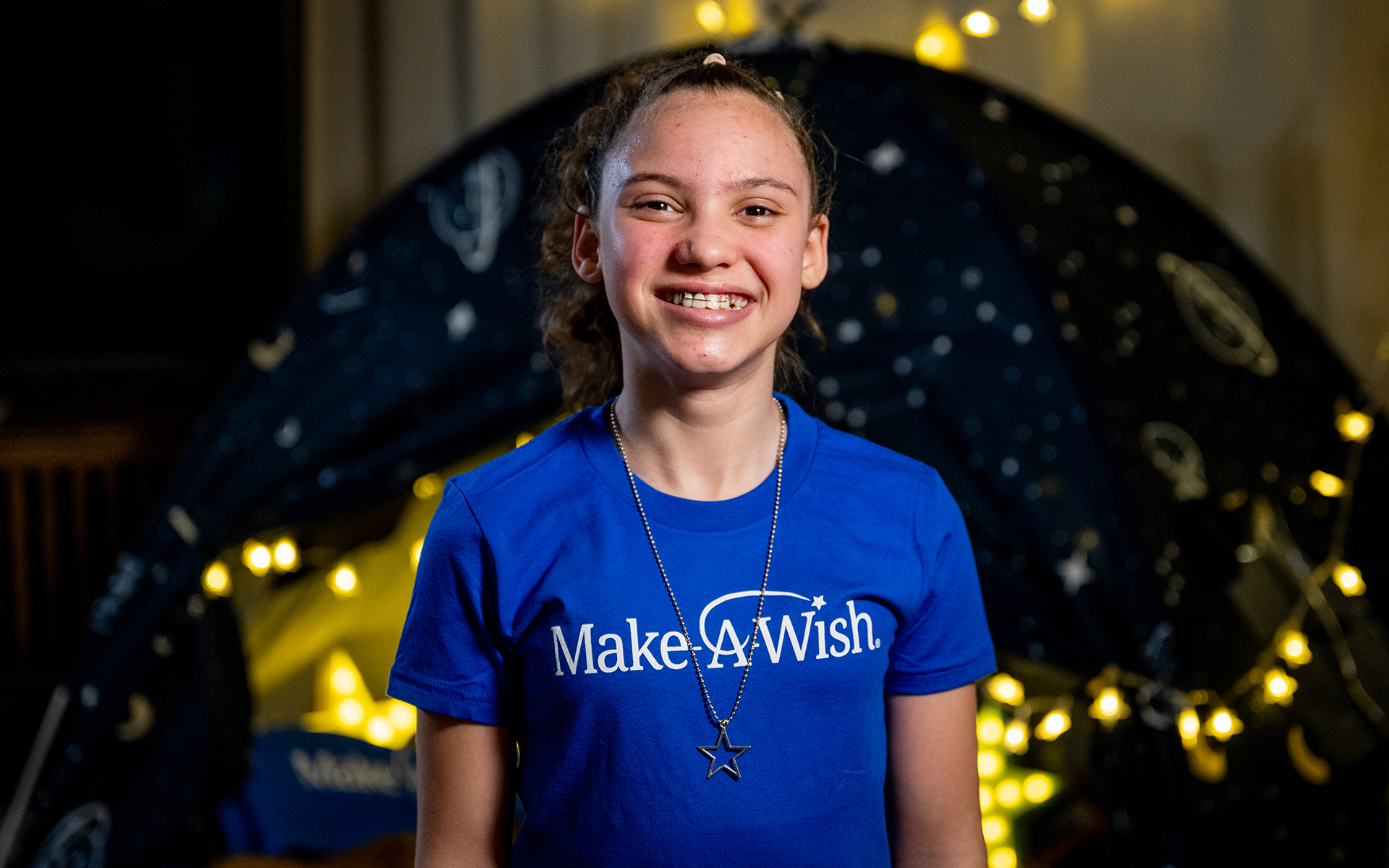 Smiling girl in Make-A-Wish shirt standing in front of tent.