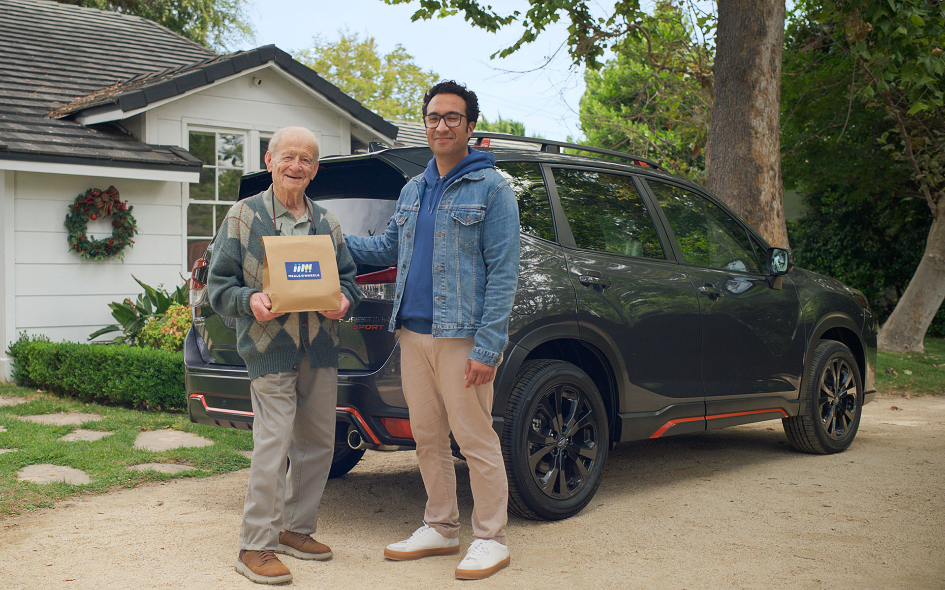 Meals on Wheels volunteer standing with senior in front of Subaru Forester and house.