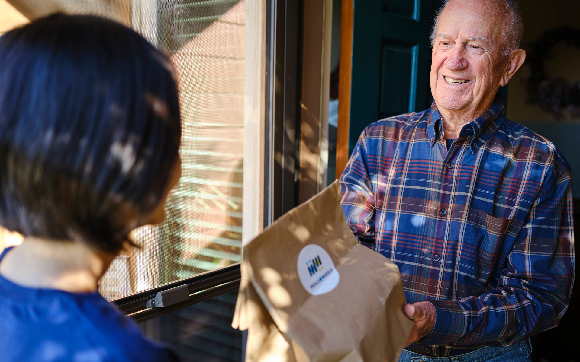 Woman in Meals on Wheels shirt delivering food to elderly man at his front door.